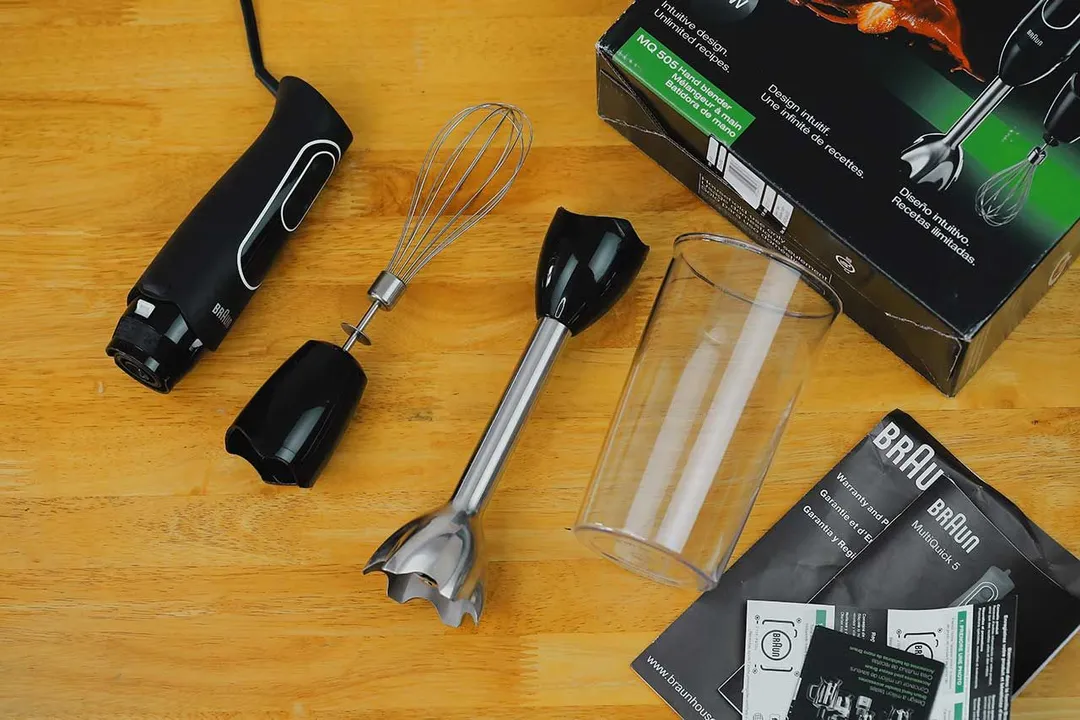The package, motor body, blending shaft, plastic beaker, user manual, and whisk attachment of the Braun MultiQuick-5 Immersion Blender on a yellow table.