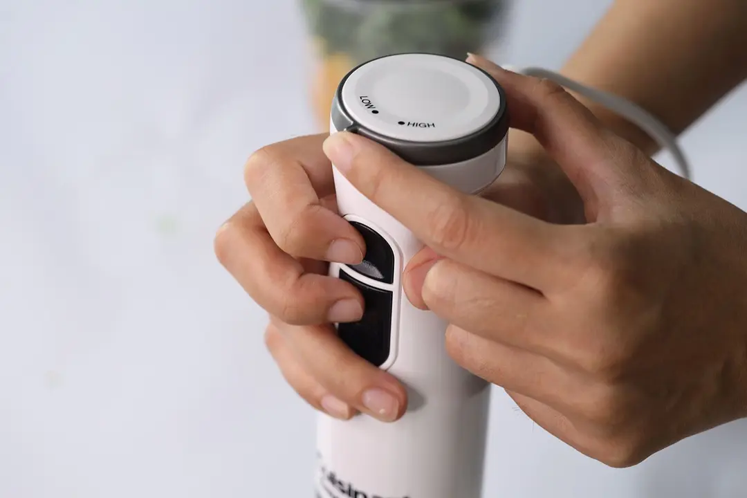 Someone uses two hands to control the Cuisinart Smart Stick immersion blender: one for holding the handle and pressing the controlling buttons at the same time, the other for rotating the speed dial. 