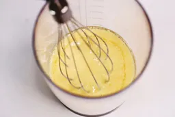 The whisk attachment of the Mueller is immersed in a plastic beaker containing egg yolks.