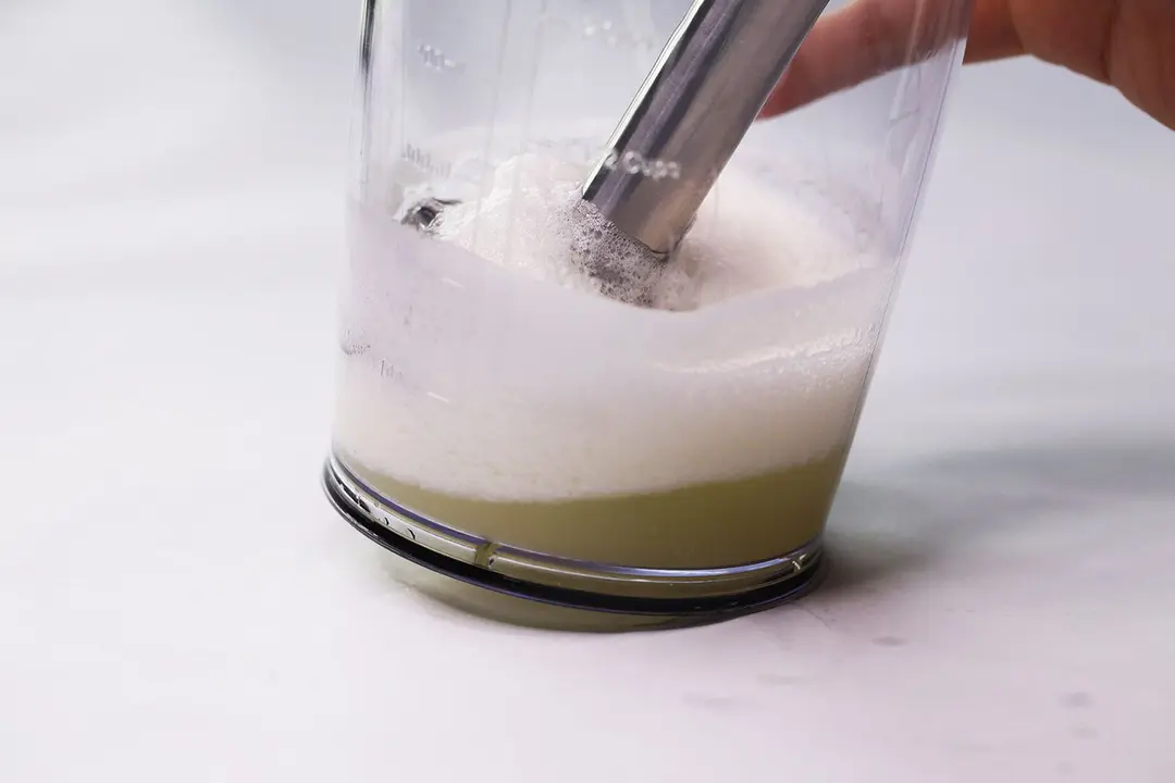 The blending wand of the KitchenAid KHBBV53 is immersed in a plastic beaker on a white table containing its failed beaten egg-white.