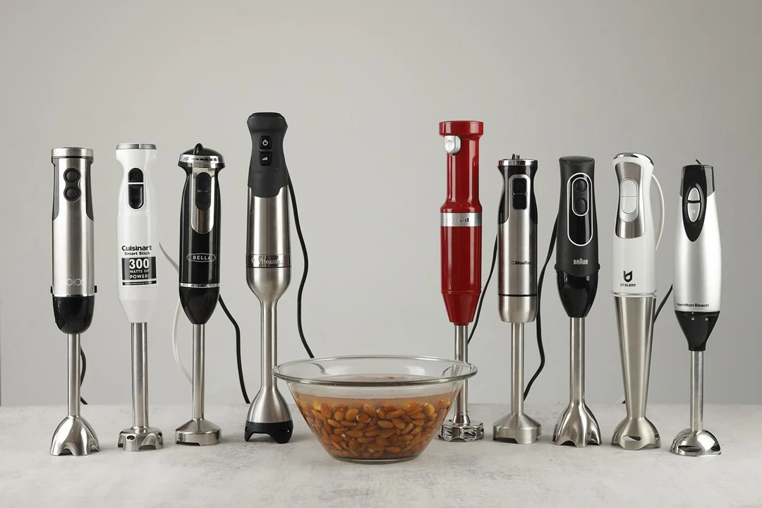 Nine immersion blenders standing on a table with a bowl of soaked almonds next to them.