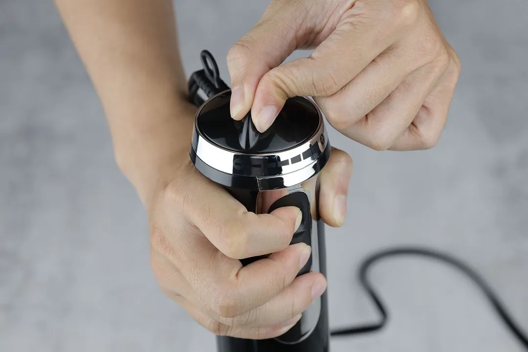 Someone uses two hands to control the BELLA immersion blender: one for holding the handle and pressing the power button at the same time, the other for rotating the speed dial. 