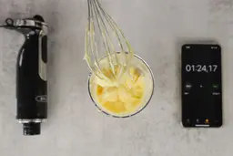A little bit of mayonnaise sticking in the whisk attachment of the BELLA hand blender when it is removed from the full batch emulsified in a plastic beaker standing between the BELLA motor body and a smartphone displaying the total emulsion time (1 minute and 24 seconds). 