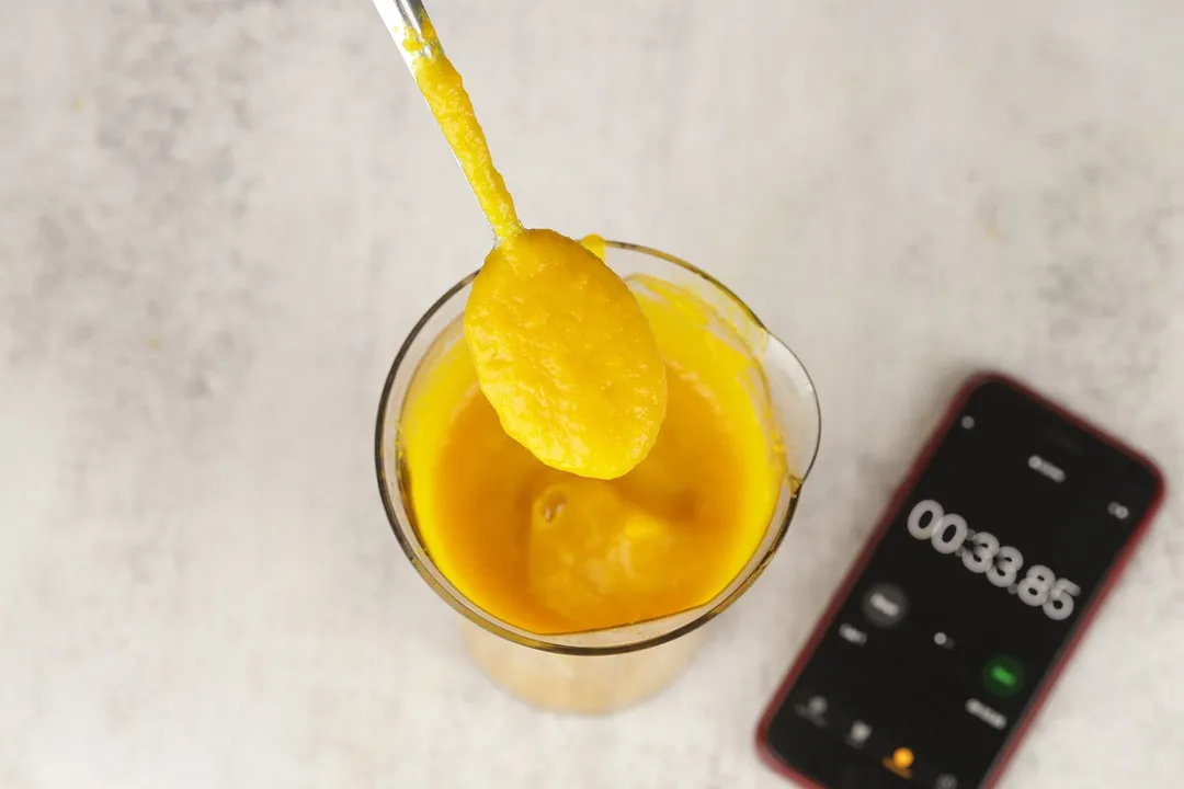Scooping a spoon of pureéd soup made by the BELLA hand blender from a plastic beaker standing next to a smartphone displaying the total pureéing time (33 seconds).  