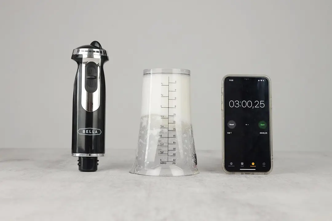 The 24-oz plastic beaker containing testing beaten egg-white is put upside down on the gray table with a BELLA motor body and a smartphone displaying the total whipping time (3 minutes) by its sides. 