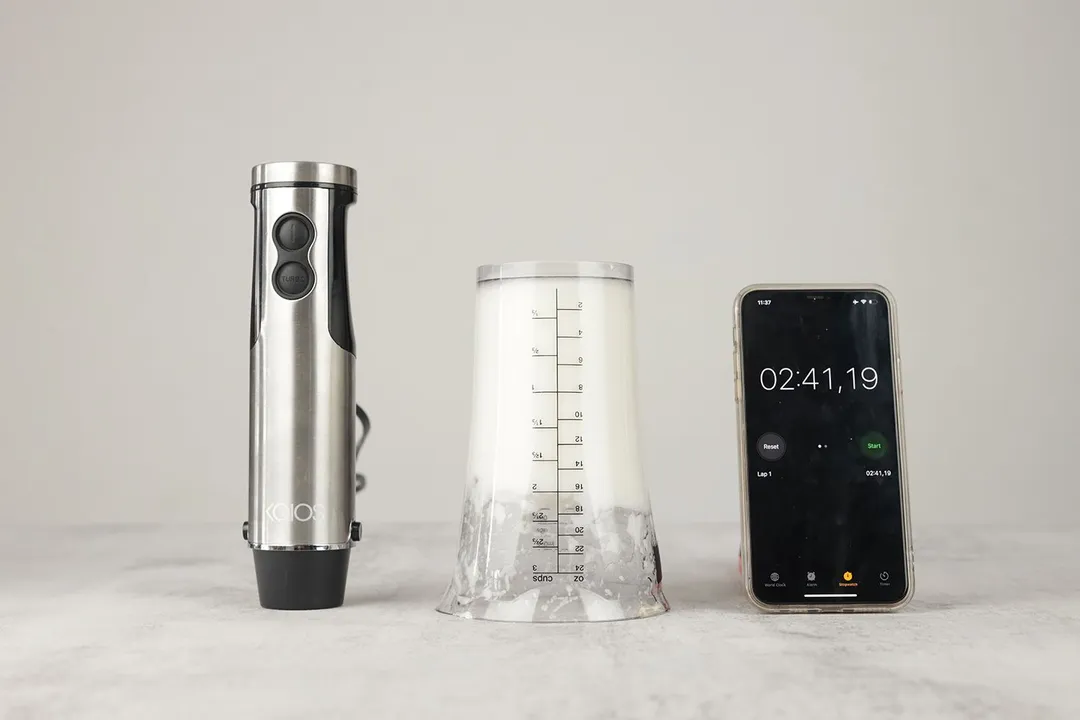 The 24-oz plastic beaker containing testing beaten egg-white of the KOIOS 4-in-1 immersion blender is put upside down on the gray table with its motor body and a smartphone displaying the total whipping time (2 minutes and 41 seconds) by its sides. 