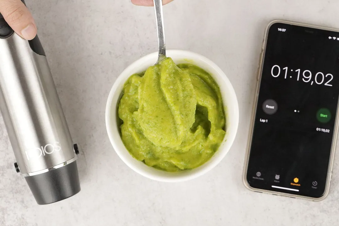 A white bowl containing a batch of green smoothie whose parts are scooped with a stainless steel spoon is between the KOIOS’s motor body and a smartphone displaying the total blending time (1 minute and 19 seconds).