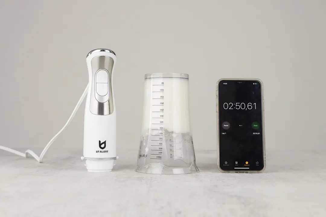 The plastic beaker containing testing beaten egg-white of the UTALENT immersion blender is put upside down on a gray table with its motor body and a smartphone displaying the total whipping time (2 minutes and 50 seconds) by its sides. 