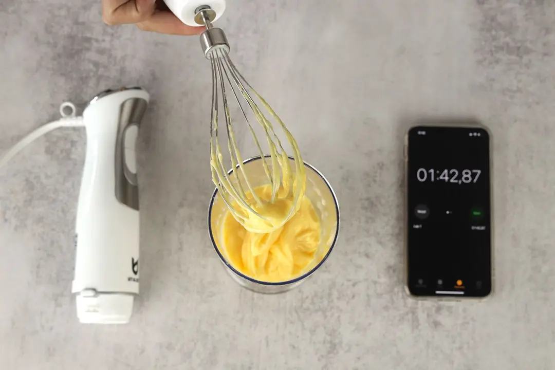 A little bit of mayonnaise sticking in the whisk attachment of the UTALENT hand blender when it is removed from the full batch emulsified in a plastic beaker standing between the motor body and a smartphone displaying the total emulsion time (1 minute and 42 seconds).