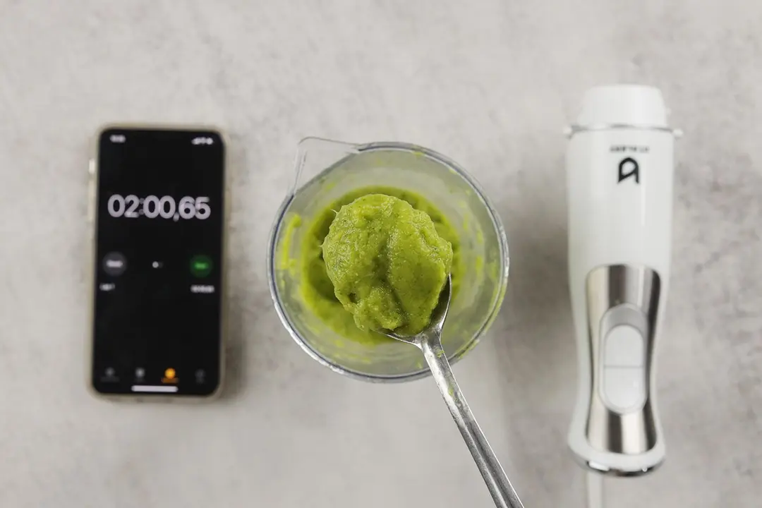 A plastic beaker containing a batch of green smoothie whose parts are scooped with a stainless steel spoon is between the UTALENT’s motor body and a smartphone displaying the total blending time (2 minutes). 
