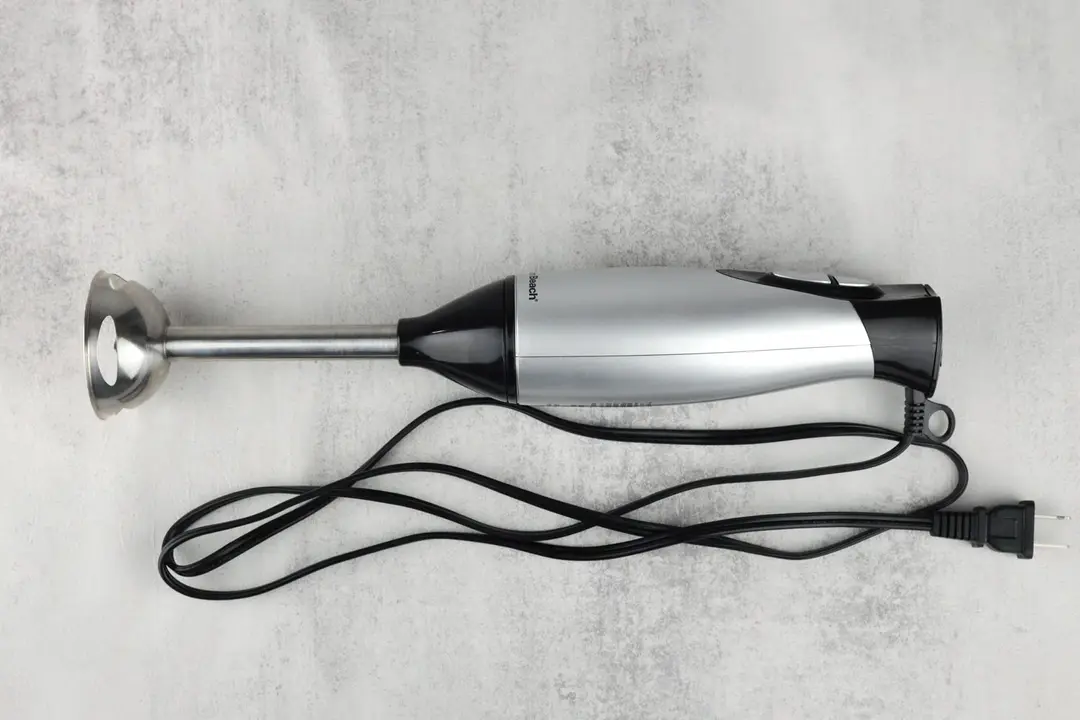 The Hamilton Beach Immersion Blender on a white table with its power cord that features a 2-prong plug rolled up next to it.