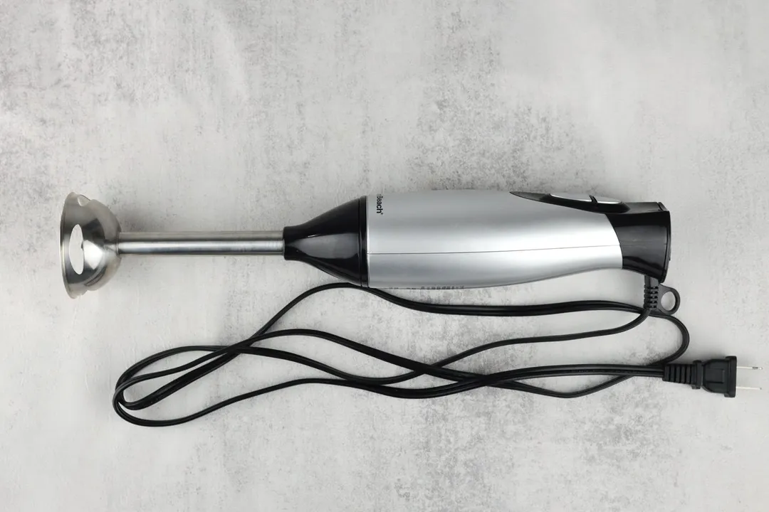 The Hamilton Beach Immersion Blender on a white table with its power cord that features a 2-prong plug rolled up next to it.