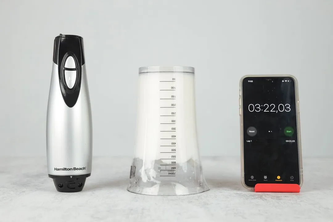 The plastic beaker containing testing beaten egg-white of the Hamilton Beach immersion blender is put upside down on the gray table with its motor body and a smartphone displaying the total whipping time (3 minutes and 22 seconds) by its sides.