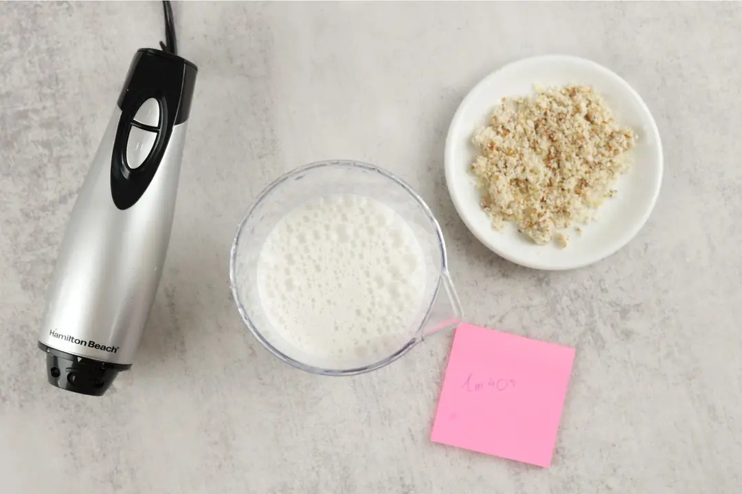 The Hamilton Beach motor body, a plastic beaker containing almond milk, a white plate of almond pulp, and a small red note displaying the total grinding time (1 minute 40 seconds) being side by side. 