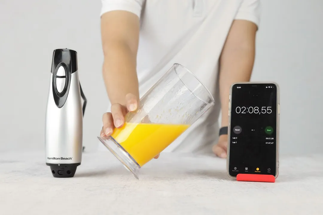 Someone is holding a plastic beaker which contains a batch of failed mayonnaise of the Hamilton Beach immersion blender and is between the motor body and a smartphone displaying the total emulsifying time (2 minutes and 8 seconds).