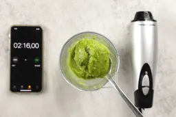 A plastic beaker containing a batch of green smoothie whose parts are scooped with a stainless steel spoon is between the Hamilton Beach motor body and a smartphone displaying the total blending time (2 minutes and 16 seconds).