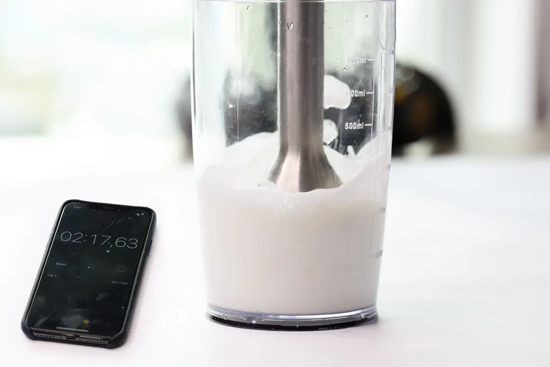 The Vitamix blending wand is immersed in a plastic beaker containing failed beaten egg-white with a smartphone displaying the total beating time ( 2 minutes and 17 seconds) next to it. 