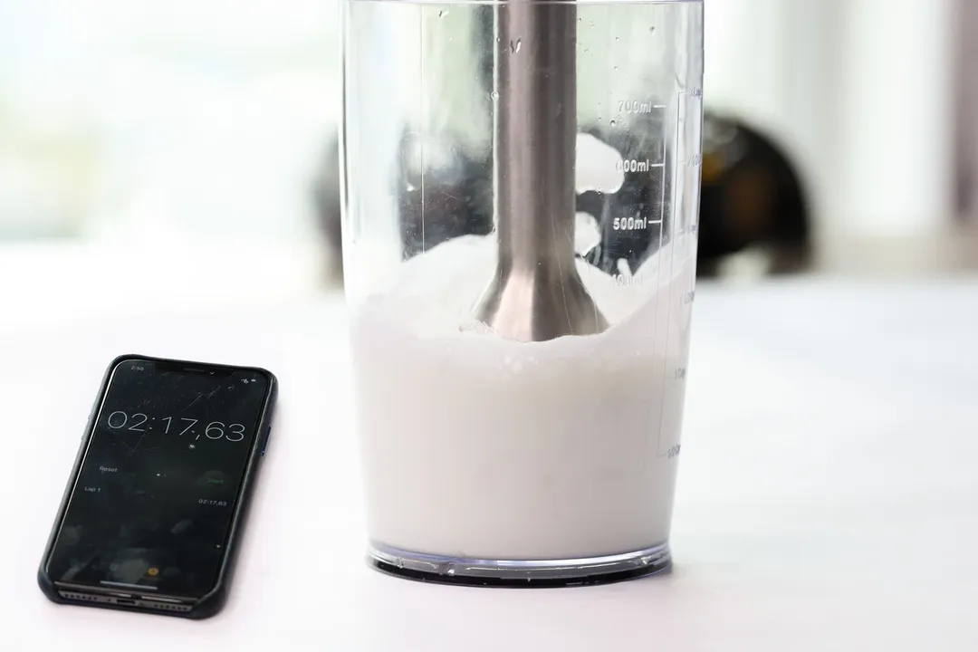 The Vitamix blending wand is immersed in a plastic beaker containing failed beaten egg-white with a smartphone displaying the total beating time ( 2 minutes and 17 seconds) next to it. 