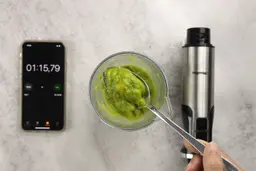 A plastic beaker containing a batch of green smoothie whose parts are scooped with a stainless steel spoon is between the Mueller’s motor body and a smartphone displaying the total blending time (1 minute and 15 seconds).