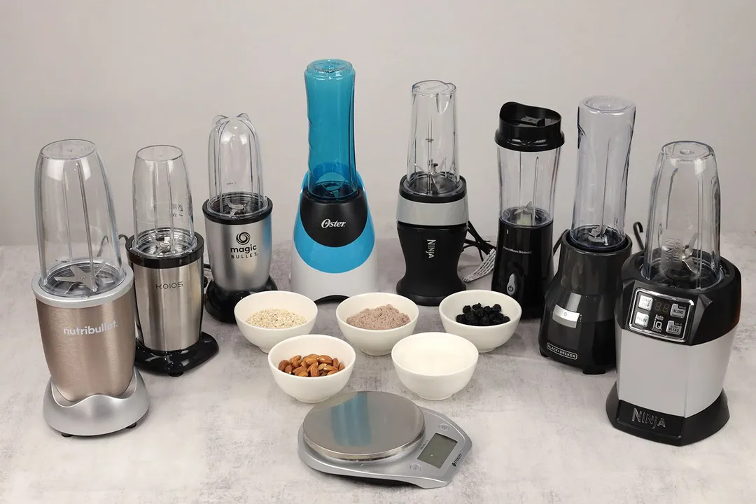 Eight personal blenders standing on a table with a scale and 5 bowls of ingredients, including protein powder, oatmeal, almonds, dried blueberries, and whole milk, next to them.
