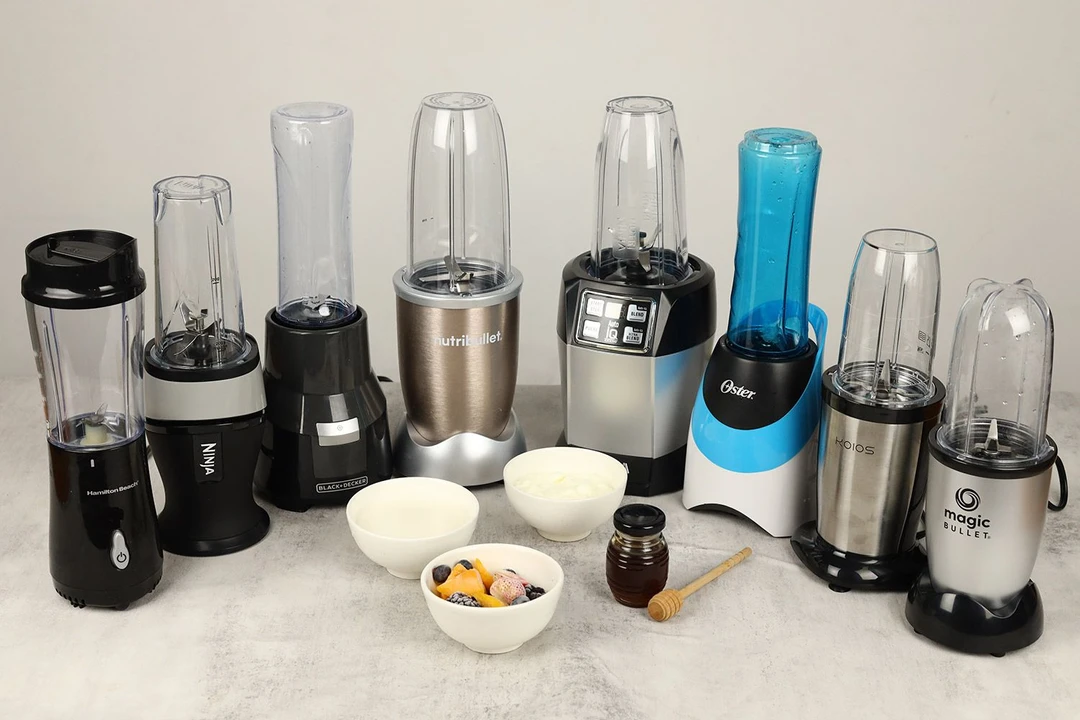 Eight personal blenders standing on a table with all the ingredients for the frozen fruit smoothie test, including yogurt, honey, whole milk, frozen fruits (mango, strawberries, blueberries, and blackberries) next to them. 