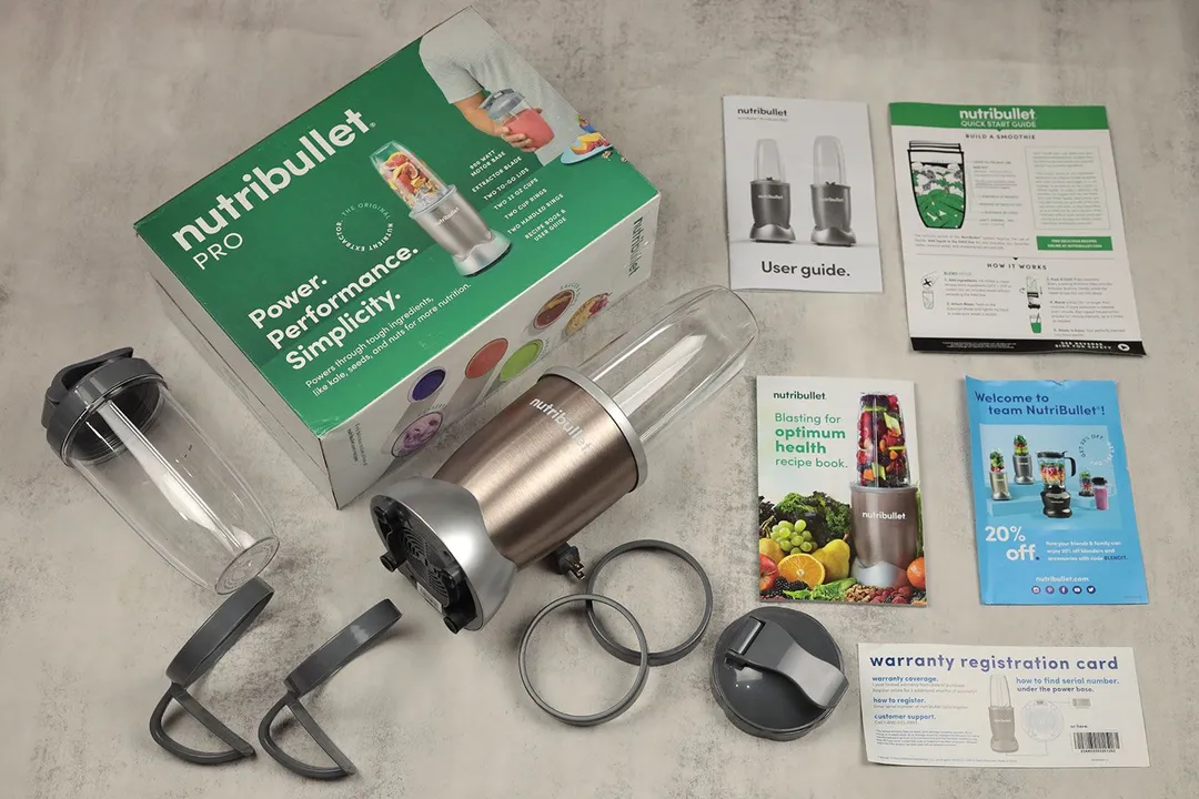 The NutriBullet Pro 900-watt personal blender lying on a gray table with its accessories by its side, including two blending cups, two to-go lids, two handled cup rings, two cup rings, a paper carton box, a recipe book, and owner’s manuals.