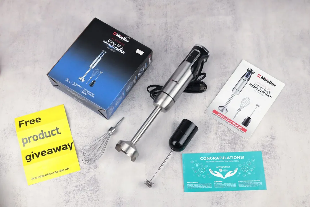 The Mueller Immersion Blender milk frother attachment, motor body, blending wand, whisk attachment, paper carton box, and user manuals lying side by side on a table.