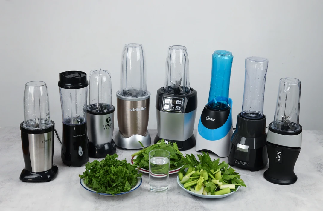 Eight personal blenders standing on a table with a cup of water and three plates of fibrous greens, including kale, spinach, and celery, next to them.