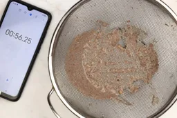 A batch of protein shake packed with dried blueberries, oatmeal, and almonds prepared by the KOIOS Bullet Personal Blender is checked for smoothness by being drained through a stainless steel mesh strainer, with a smartphone displaying the total blending time (56 seconds) next to it. 