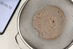 A batch of protein shake prepared by the Ninja Small Blender is checked for smoothness by being drained through a stainless steel mesh strainer, with a smartphone displaying the total blending time (50 seconds) next to it.
