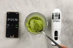 A plastic beaker containing a batch of green smoothie whose parts are scooped with a stainless steel spoon is between the Cuisinart’s motor body and a smartphone displaying the total blending time (1 minute and 21 seconds).