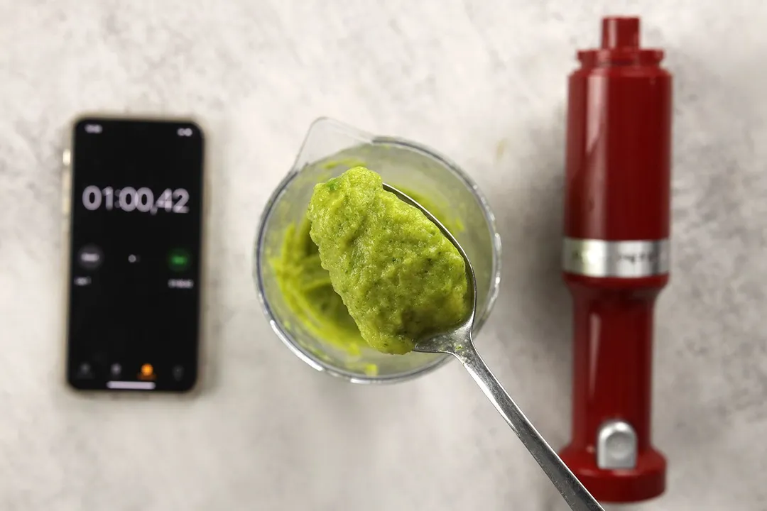 A plastic beaker containing a batch of green smoothie whose parts are scooped with a spoon is standing between a timer displaying the total blending time (1 minute) and the KitcheAid’s motor body. 