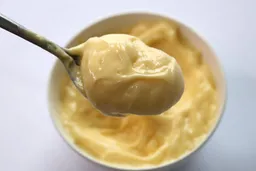 Scooping a spoon of mayonnaise emulsified by the KitchenAid immersion blender from a white bowl to check its creaminess. 