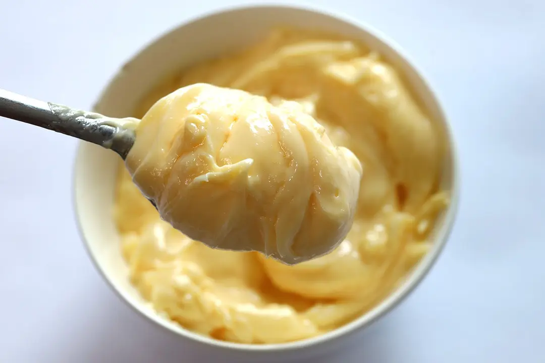 Scooping a spoon of mayonnaise emulsified by the Vitamix immersion blender from a white bowl to check its creaminess. 