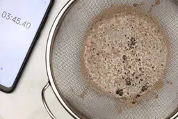A batch of protein shake prepared by the Hamilton Beach Single-Serve Blender is checked for smoothness by being drained through a stainless steel mesh strainer, with a smartphone displaying the total blending time (3 minutes and 45 seconds) next to it. 