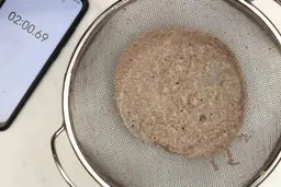 A batch of protein shake prepared by the Black and Decker PB1002 blender is checked for smoothness by being drained through a stainless steel mesh strainer, with a smartphone displaying the total blending time (2 minutes) next to it.