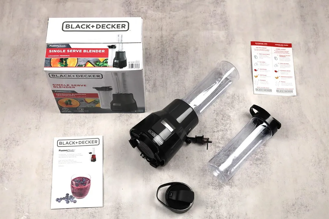 The Black+Decker Fusionblade lying on a table with its additional accessories, including an extra blending cup with lid, a to-go lid, a paper carton box, and owner’s manuals.