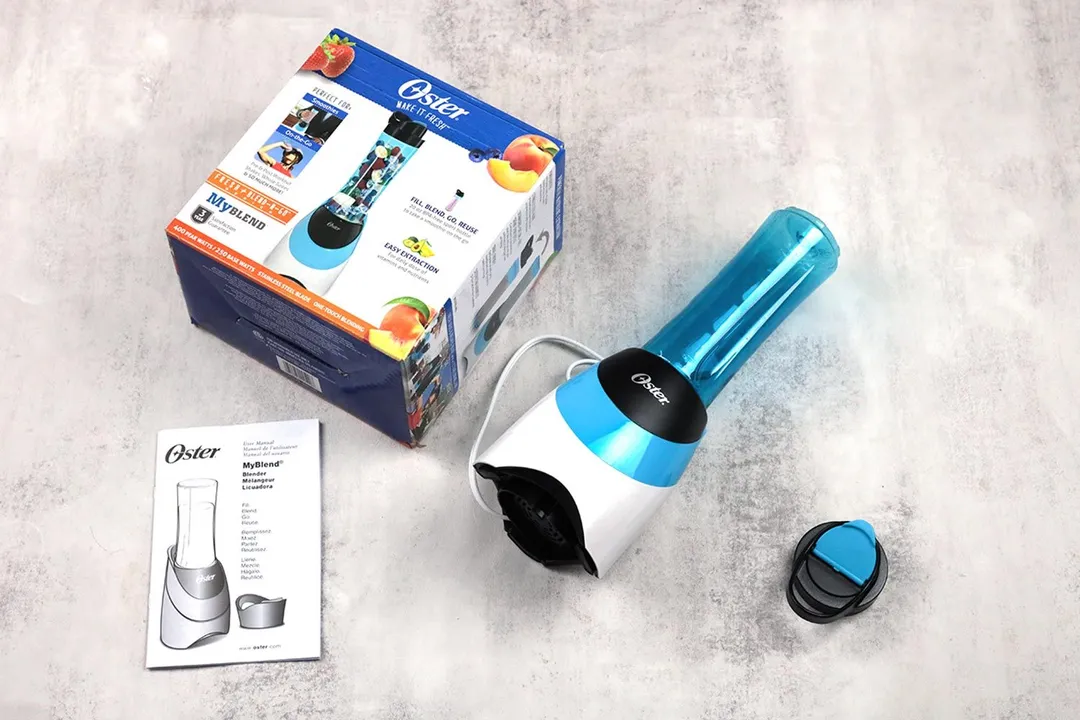 The Oster Myblend personal blender lying on a table with its additional accessories, including a to-go lid, user guide, and paper carton box.