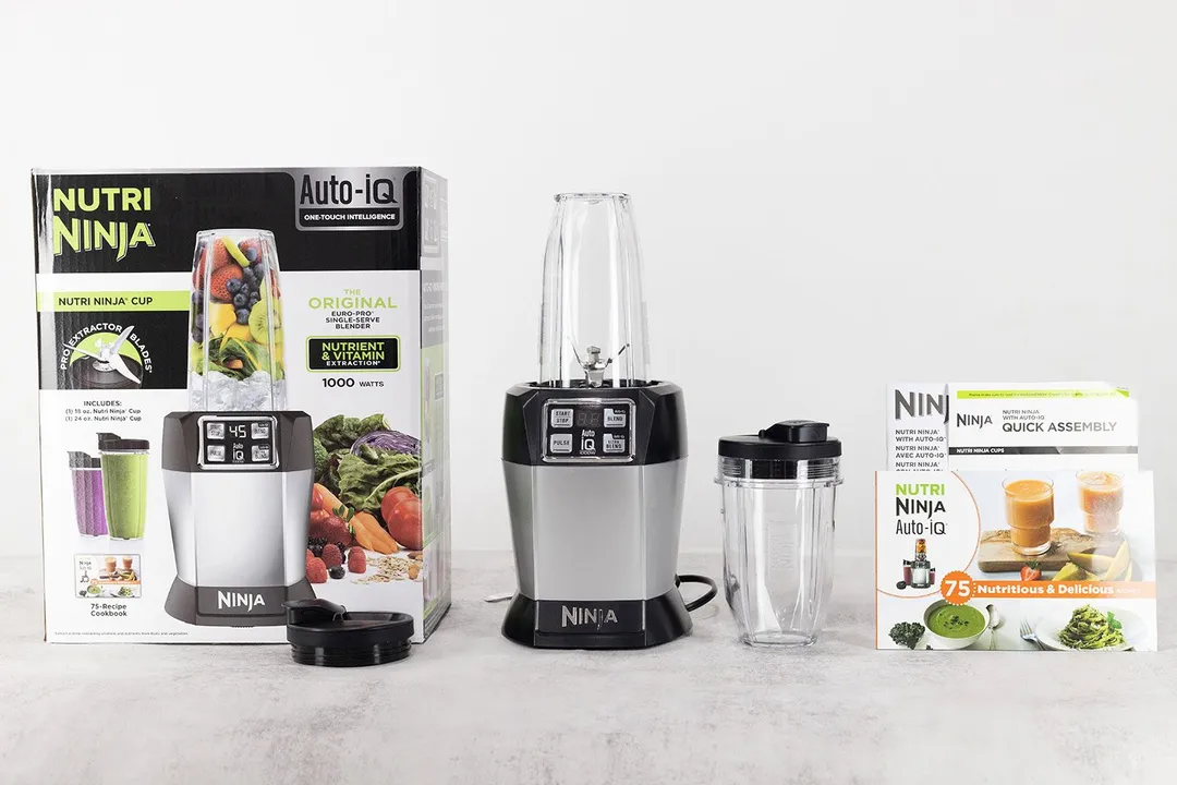 The Ninja Nutri Auto-IQ personal blender 1000-Watt standing on a gray table with its accessories, including two to-go lids, an additional blending cup, a recipe book & user guide, and a paper carton box, by its sides. 