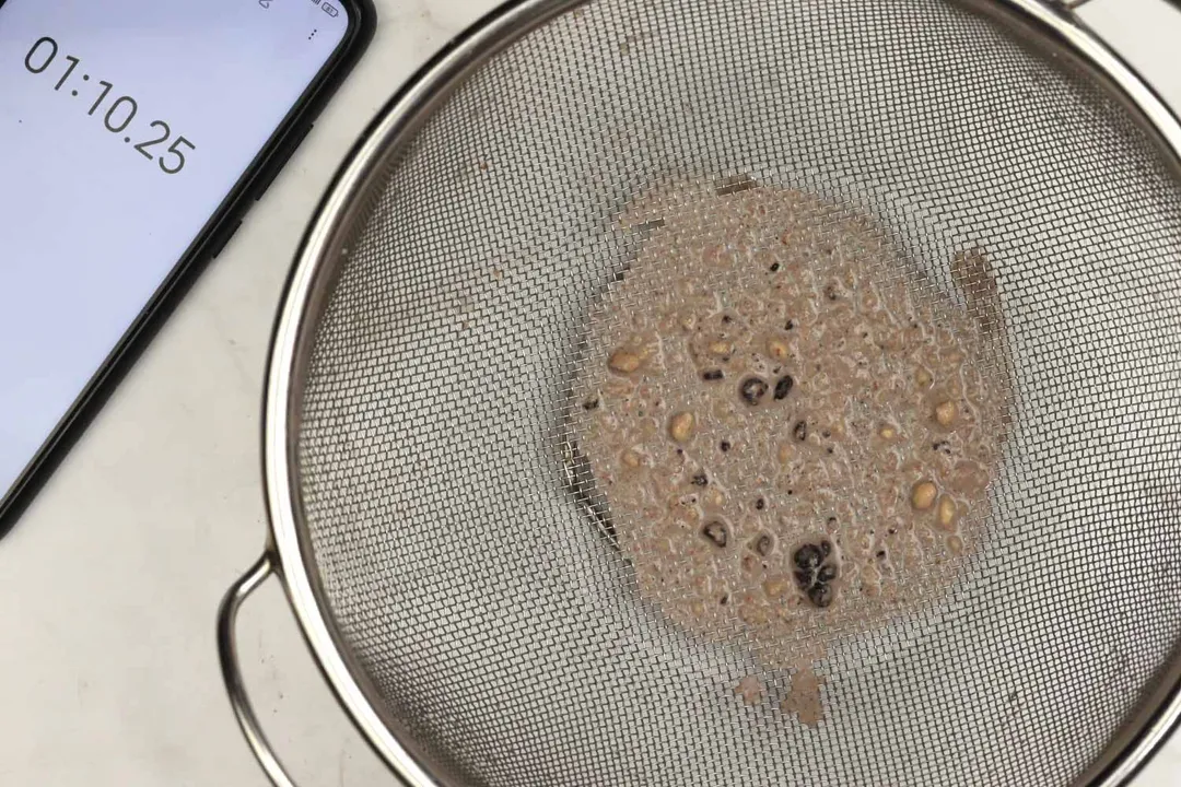 A batch of protein shake packed with dried blueberries, oatmeal, and almonds prepared by the Ninja BL480D is checked for smoothness by being drained through a stainless steel mesh strainer, with a smartphone displaying the total blending time (1 minute and 10 seconds) next to it. 