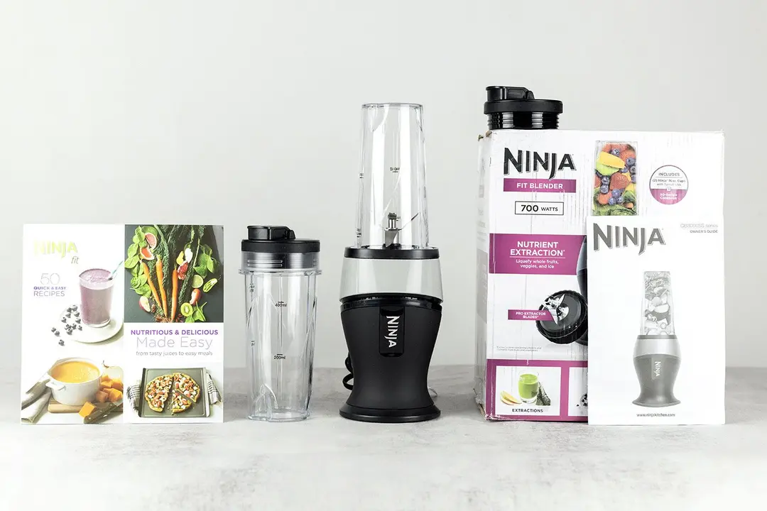 The Ninja Fit personal blender 700-Watt personal blender standing on a gray table with its additional accessories by its side, including an extra blending cup, two to-go lids, a user’s manual, and a paper carton box. 