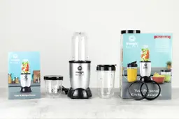 The Magic Bullet 250W Personal Blender and its additional accessories by its side, including a to-go lid, two additional blending cups, a party mug, two lip rings, two resealable lids, a paper carton box, and a user & recipe book guide.