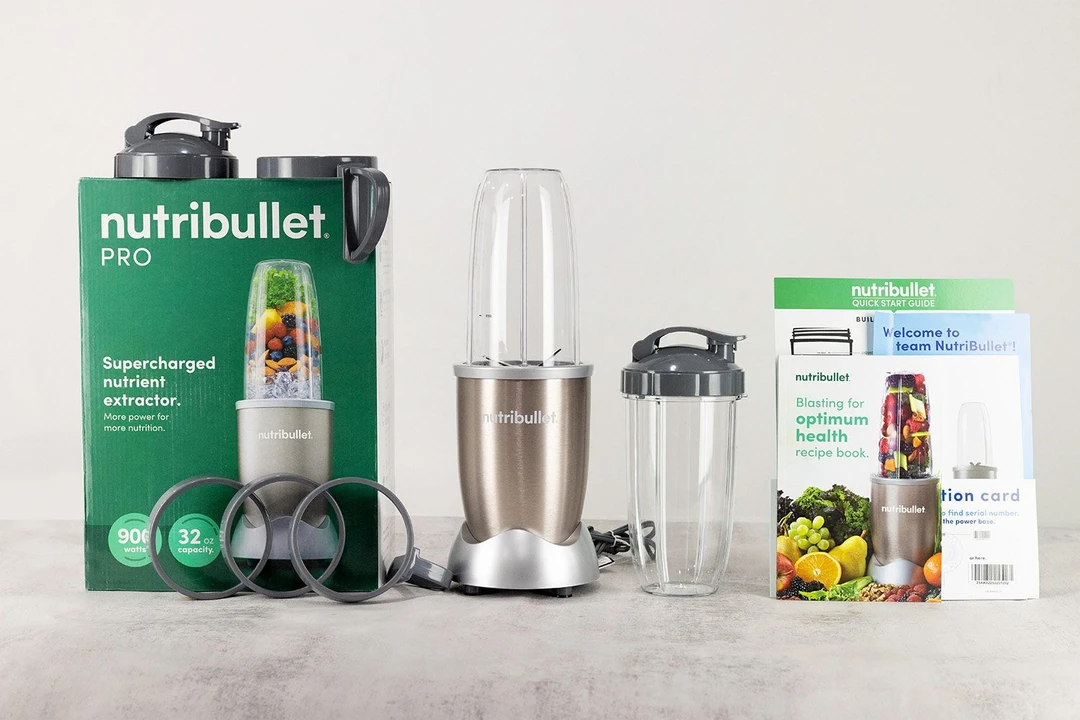 The NutriBullet Pro 900 Personal Blender and its additional accessories by its side, including  two to-go lids, two blending cups, two cup rings, two handled cup rings, a paper carton box, a recipe book, and a user guide.