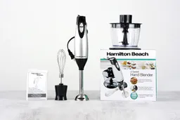 The Hamilton Beach hand blender user’s manual, together with a whisk attachment, a blending wand with its motor body attached, and a paper carton box with a food processor attachment above, standing on a gray table.