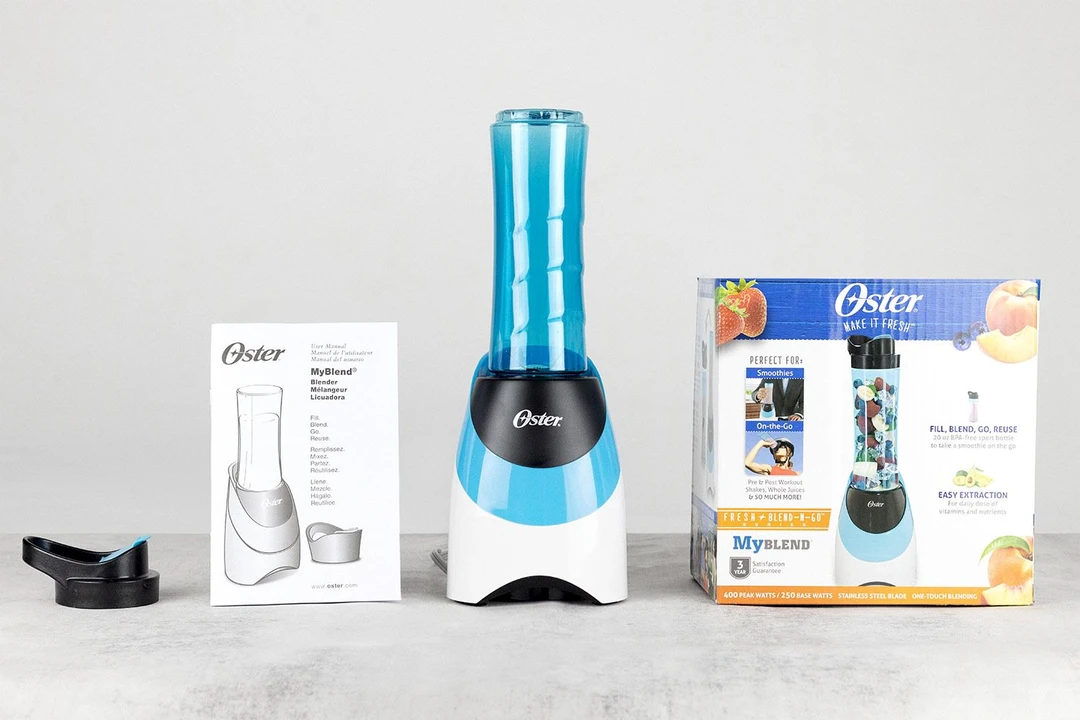 The Oster personal blender standing on a white table with its additional accessories, including a to-go lid, user’s manual, and paper carton box, by its sides.