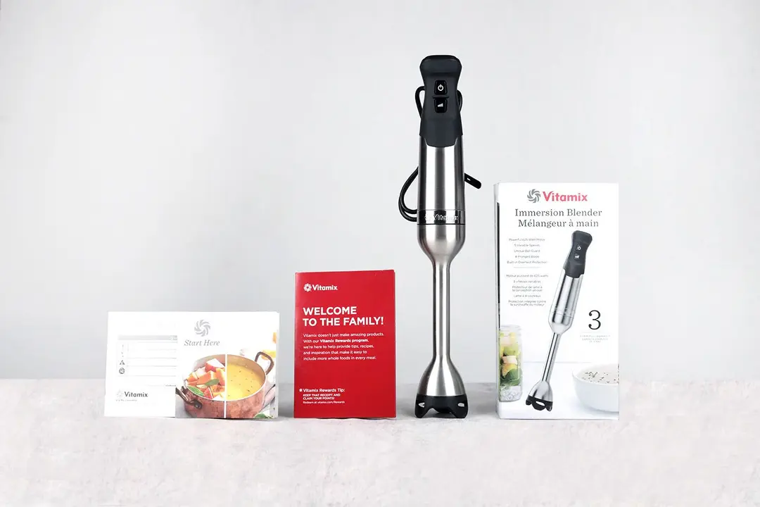 Unboxing the Vitamix immersion blender, from left to right: an owner’s manual, a recipes booklet, a red receipt, the Vitamix 5-speed with a stainless steel motor body and an immersion blending wand, and a paper carton box.