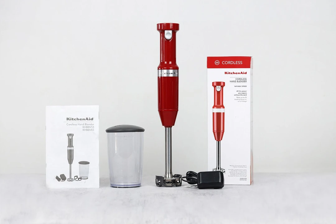 Unboxing the KitchenAid Cordless Immersion BLender, from left to right: an user manual, a beaker and lid, the KitchenAid KHBBV53 with its motor body and blending wand, a charger, and a paper carton box.
