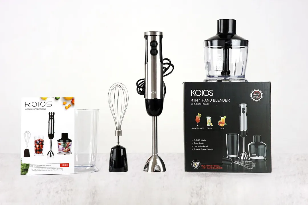 The koios 4 in 1 hand blender standing on the gray table with its accessories, including a user manual, plastic beaker, whisk attachment, food processor located above a paper carton box. The KOIOS 4-in-1 immersion blender standing on the gray table with its accessories, including a user manual, plastic beaker, whisk attachment, food processor located above a paper carton box.