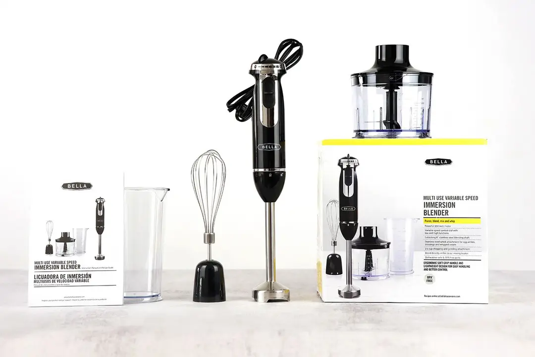 The BELLA immersion blender standing on the gray table with its accessories, including a user manual, plastic beaker, whisk attachment, food processor located above a paper carton box.