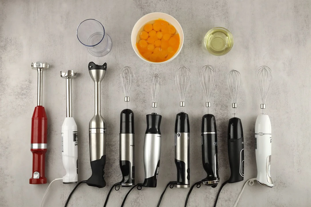 Nine immersion blenders lying on a table with ingredients and necessary tools for the mayonaise test, including a plastic beaker, a bowl of egg yolk, and a cup of oil, next to them.